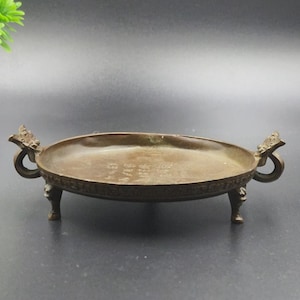 May include: A bronze-colored, oval-shaped tray with ornate handles and legs. The tray has a slightly raised rim and is supported by three small, decorative legs. The handles feature a curved design. The tray appears to be made of metal.
