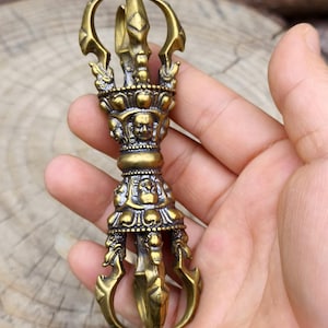 Könnte beinhalten: Ein detaillierter Vajra aus Messing, ein Ritualobjekt mit einem zentralen Dorn und gebogenen Klingen. Das Stück im antiken Stil weist aufwendige Schnitzereien von Gesichtern und verzierten Mustern auf. Das Objekt wird in einer Hand gehalten, um seine Größe und sein Design zu zeigen.