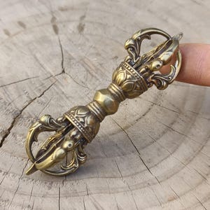 Könnte beinhalten: Ein detaillierter Vajra in Bronzefarbe, ein rituelles Objekt, mit komplizierten floralen und geometrischen Mustern. Das Objekt hat einen zentralen, bauchigen Abschnitt und gebogene Zacken an jedem Ende. Der Vajra wird von einem Finger gehalten, der seine Größe und Details zeigt.