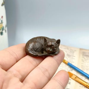 May include: A small, bronze-colored cat figurine in a curled-up sleeping position. The cat is resting on a hand, with visible details of its fur and facial features. The figurine is a miniature sculpture, likely for decorative purposes.