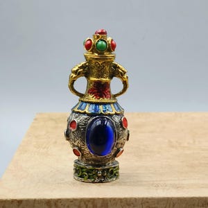 May include: An ornate, gold-coloured perfume bottle with a crown-like top, featuring red and green jewels. The bottle has a blue oval gem and is decorated with silver and red accents. The base is green with a floral design.