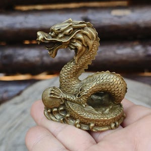 May include: A detailed brass dragon figurine, posed with an open mouth and a ball. The dragon has a scaled body and a detailed head. The figurine is a golden-brown color and appears to be made of metal.