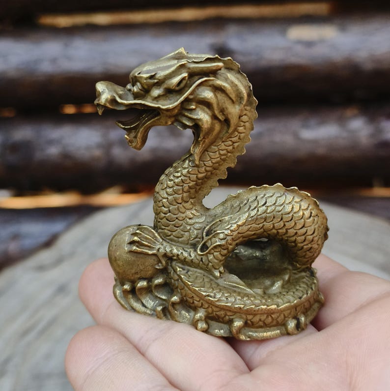 May include: A detailed brass dragon figurine, posed with an open mouth and a ball. The dragon has a scaled body and a detailed head. The figurine is a golden-brown color and appears to be made of metal.