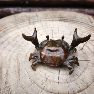 May include: A bronze crab figurine with detailed claws and eyes. The crab is positioned on a light-colored, circular wooden surface, showcasing the texture of the wood grain. The crab's body has a textured surface.