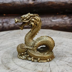May include: A detailed brass dragon figurine with an open mouth and intricate scale details. The dragon is posed on a round base, with a ball held in its claw. The figurine is approximately 4 inches tall.