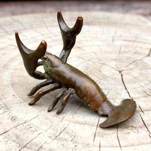 Puede incluir: Una pequeña escultura metálica de color bronce que representa una langosta o un cangrejo de río. La figura presenta dos pinzas abiertas y elevadas, varias patas articuladas y un cuerpo segmentado con una cola ancha y plana, con pátina.
