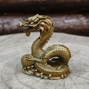 May include: A detailed brass dragon figurine with an open mouth and scaled body. The dragon is posed in a curved position, with its tail curled around its base. The figurine is a golden color and sits on a wooden surface.