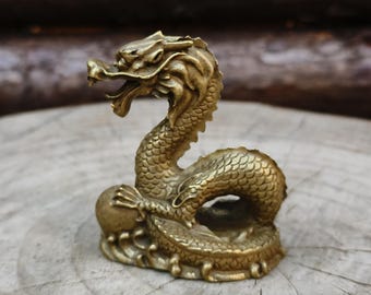 May include: A detailed brass dragon figurine with an open mouth and scaled body. The dragon is posed in a curved position, with its tail curled around its base. The figurine is a golden color and sits on a wooden surface.