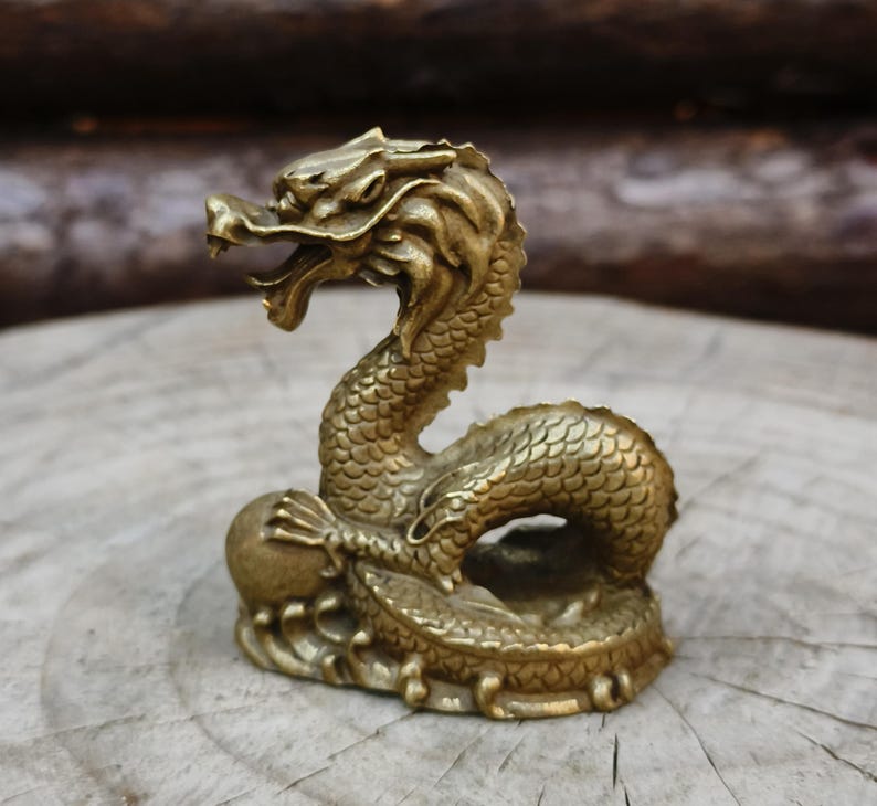 May include: A detailed brass dragon figurine with an open mouth and scaled body. The dragon is posed in a curved position, with its tail curled around its base. The figurine is a golden color and sits on a wooden surface.