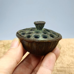 May include: A bronze-colored, lotus-shaped container with a lid. The lid has multiple circular openings. The container is held in a hand. The container has a textured surface and a small, cone-shaped handle on top.