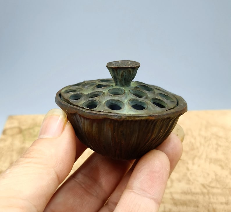May include: A bronze-colored, lotus-shaped container with a lid. The lid has multiple circular openings. The container is held in a hand. The container has a textured surface and a small, cone-shaped handle on top.