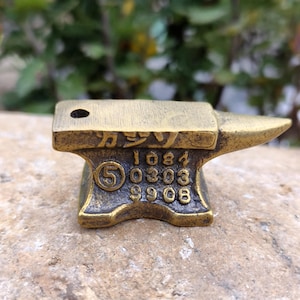 May include: A small, antique-style brass anvil with a hole on top. The anvil has a flat top and a pointed horn. The base features the number 5 and several other numbers. Japanese characters are etched on the top.