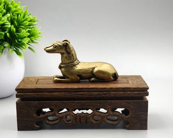 May include: A golden-colored dog figurine resting on a dark brown wooden display stand. The dog is in a relaxed pose, with its legs tucked beneath it. The stand has a decorative, carved design.