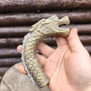 Könnte beinhalten: Eine detaillierte Drachenkopfskulptur im antiken Stil in messinggoldener Farbe. Der Drachenkopf ist offenmäulig, mit komplizierten Schuppen- und Gratdetails. Die Skulptur ist gebogen und scheint ein Griff oder ein dekoratives Element zu sein.