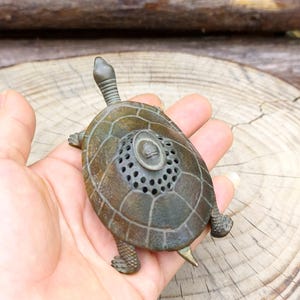 Peut inclure: Figurine de tortue de couleur bronze avec un motif de carapace détaillé et une petite tête relevée. La tortue a de petits pieds et une queue. La carapace présente un motif circulaire avec de petits trous. La figurine est tenue dans une main.