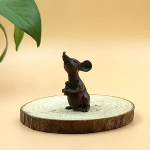 Könnte beinhalten: Eine bronzefarbene Mäusefigur mit nach oben geneigtem Kopf, die ein kleines Objekt hält. Die Maus steht auf einer runden Holzscheibe mit natürlichem Rindenrand. Der Hintergrund ist einfarbig hellbraun.
