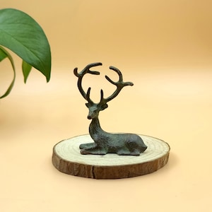 May include: A bronze-coloured deer figurine in a resting pose, mounted on a round, natural wood slice. The deer has detailed antlers and is set against a soft, yellow background. A decorative item.