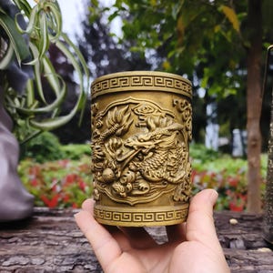 May include: A cylindrical, antique gold-colored container with intricate dragon carvings. The container features a repeating geometric pattern at the top and bottom. The dragon design is highly detailed, showcasing scales, claws, and a dynamic pose. The container is held in a hand.