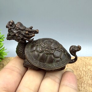 May include: A bronze-coloured figurine of a mythical dragon-turtle hybrid. The sculpture has intricate details, including a dragon's head and a turtle's shell. The tail is curled. The figurine is held in a hand.