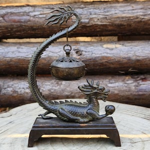 Könnte beinhalten: Bronzener Drachen-Räuchergefäß. Der geschwungene Schwanz trägt eine hängende Schale mit Deckel. Der Drache ruht auf einem rechteckigen Sockel und hält eine Kugel. Das Räuchergefäß ist mit Schuppen und einem stilisierten Drachenkopf versehen.