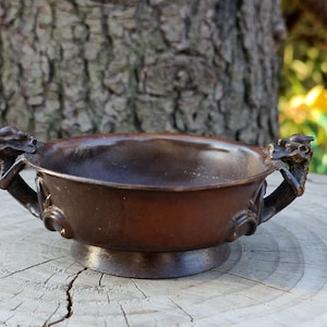 May include: A bronze-colored bowl with dragon-shaped handles. The bowl has a round base and a wide, shallow basin. The handles are intricately detailed, and the bowl appears to be made of metal. The bowl is likely an antique or decorative item.