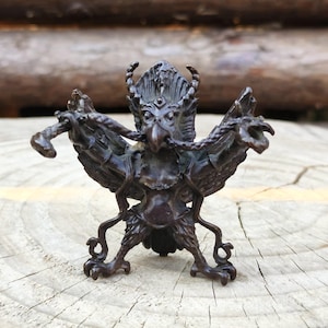 Può includere: Statuetta color bronzo di una creatura mitica con testa di uccello, ali e piedi artigliati. La figura presenta dettagli intricati, tra cui un copricapo e lunghi baffi curvi. Le ali sono spiegate.