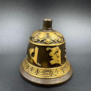 Op de afbeelding: Een bronzen bel met gouden details. De bel heeft reliëf bloempatronen en gestileerde karakters. De basis heeft een geometrisch ontwerp. De bel is waarschijnlijk voor religieus of decoratief gebruik.