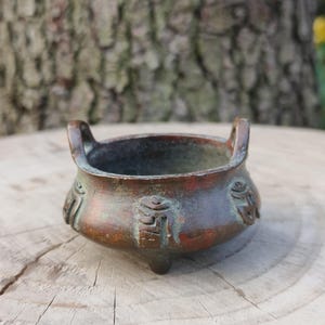 May include: A small, antique bronze incense burner with three small feet and two handles. The bowl has an aged patina with green and brown tones. Decorative symbols are etched on the sides.