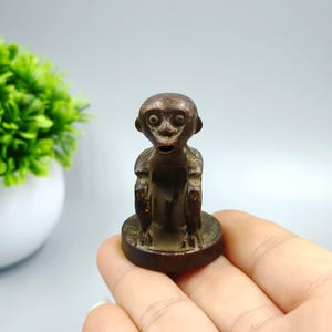 May include: A small, brown monkey figurine with a surprised expression. The monkey is seated on a round base. The figurine is held in a hand, with a green plant in a white pot in the background.