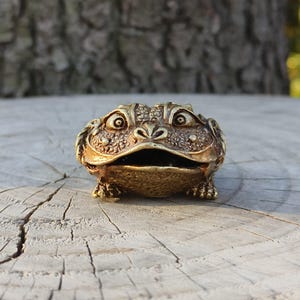 May include: A detailed, golden-colored frog figurine with an open mouth and textured skin. The frog is positioned on a wooden surface, showcasing its intricate design and craftsmanship. The figurine's eyes are wide, and its pose is lifelike.