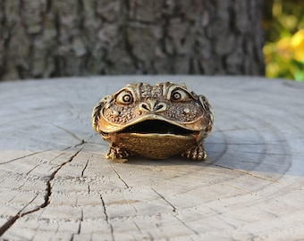 Puede incluir: Figurilla detallada de rana dorada con la boca abierta y piel texturizada. La rana está colocada sobre una superficie de madera, mostrando su intrincado diseño y artesanía. Los ojos de la figurilla son grandes.