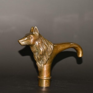 Puede incluir: Un mango de bastón de color bronce con una cabeza de lobo detallada. El pelaje del lobo es texturizado y el mango se curva suavemente. Un accesorio decorativo.