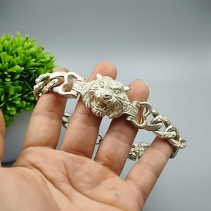 Puede incluir: Pulsera plateada con un detallado centro de cabeza de tigre. La pulsera tiene un diseño de eslabones de cadena con cierres decorativos. La cabeza de tigre está intrincadamente esculpida, mostrando los rasgos del animal.
