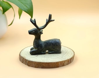 May include: A bronze-colored deer figurine with detailed antlers, resting on a round, natural wood slice. The deer is in a relaxed, seated pose. The wood slice has a rough, textured edge.
