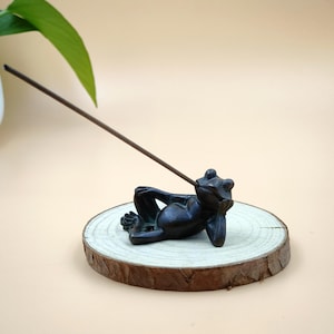 Puede incluir: Un porta incienso de rana de color bronce, reclinado sobre una rodaja de madera. La rana está en una pose relajada, sosteniendo un palito de incienso. La rodaja de madera tiene un borde natural sin terminar.