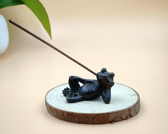 May include: A bronze-colored frog incense holder, reclining on a wooden slice. The frog is in a relaxed pose, holding an incense stick. The wooden slice has a natural, unfinished edge. The background is a soft, neutral color.