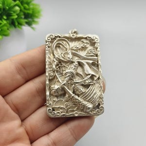 Può includere: Ciondolo rettangolare in argento con un rilievo dettagliato di una figura di guerriero, nuvole e un bastone. Il ciondolo ha un bordo ornato e un anello per una catena. Il design è intricato e il metallo ha una finitura lucida.