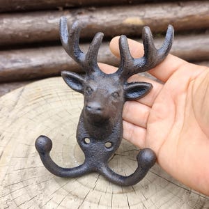 May include: A cast iron deer head wall hook with a dark, weathered brown finish. The hook has a detailed deer head with antlers and two curved hooks. Ideal for a rustic interior, it measures approximately 15 cm in height.