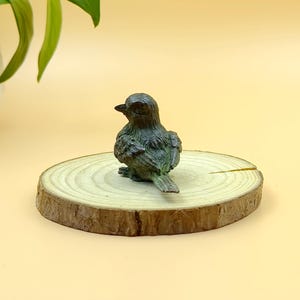 Könnte beinhalten: Eine kleine, dunkelgrüne Vogelfigur sitzt auf einer runden, natürlichen Holzscheibe. Der Vogel hat detaillierte Federn und ein leicht verwittertes Aussehen. Die Holzscheibe hat sichtbare Jahresringe und einen Rindenrand.