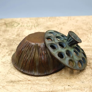 May include: A bronze-colored, bowl-shaped container with a textured exterior and a separate, perforated lid. The lid is a greenish-bronze color and has a small handle. The container and lid are placed on a textured, light-brown surface.