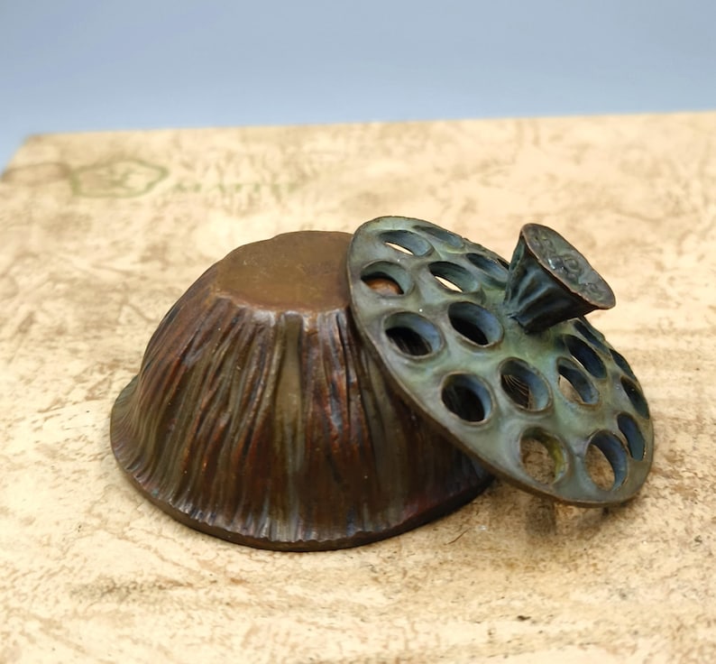 May include: A bronze-colored, bowl-shaped container with a textured exterior and a separate, perforated lid. The lid is a greenish-bronze color and has a small handle. The container and lid are placed on a textured, light-brown surface.