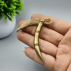 Puede incluir: Un pequeño colgante de hoz de latón de estilo antiguo. La hoja curva y el mango están detallados con una textura similar al bambú. El colgante se sostiene en una mano, mostrando su tamaño y diseño.