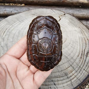 Puede incluir: Un caparazón de tortuga de madera tallada de color marrón oscuro con detalles intrincados. El caparazón presenta caracteres chinos grabados y un símbolo central de Yin Yang. El caparazón de tortuga se sostiene en una mano sobre un fondo de madera.