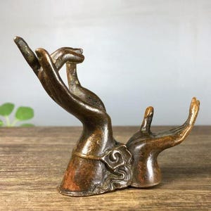 May include: Bronze sculpture depicting two hands in a gesture of offering or receiving. The hands are detailed with visible fingers and a textured surface, suggesting an antique or vintage piece. The sculpture is set on a wooden surface.