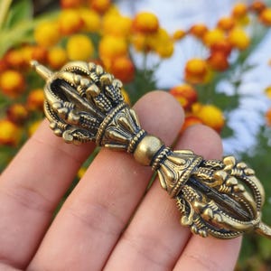 May include: A bronze-coloured vajra, a ritual object, is held against a backdrop of yellow and orange flowers. The vajra features intricate details and a symmetrical design, with ornate floral patterns.