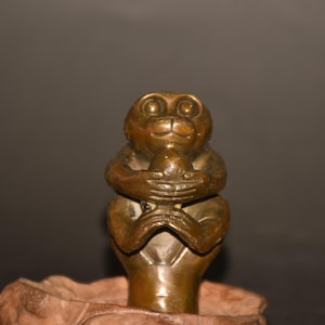 Könnte beinhalten: Eine bronzene Affenfigur, die einen kleinen Gegenstand, möglicherweise eine Frucht oder Nuss, hält. Der Affe sitzt, die Arme um den Gegenstand gelegt. Die Skulptur hat eine detaillierte, strukturierte Oberfläche und einen neutralen Hintergrund.