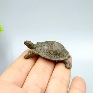 Op de afbeelding: Een kleine, bronzen schildpadfiguur. De schildpad heeft een gedetailleerd schild met een geometrisch patroon en staat met uitgestrekte poten. Het beeldje rust op een hand, wat de grootte en details laat zien.