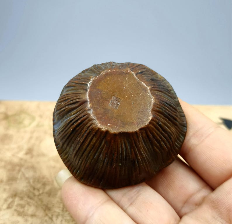 May include: A small, round, antique-style brown bowl with a textured, ridged exterior. The bowl's interior is a lighter brown with a stamped design. The bowl is held in a person's hand, with a neutral background.
