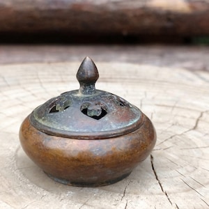 May include: A small, antique bronze incense burner. It has a rounded base and a perforated lid with heart-shaped cutouts and a pointed finial. The burner has a weathered appearance, suggesting it is vintage or handcrafted. The measurements are in cm.