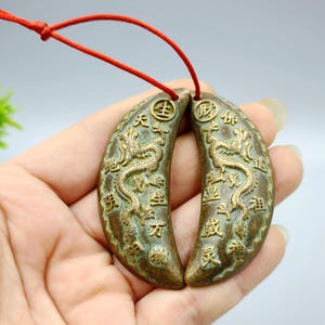 May include: Two bronze-colored, crescent-shaped pendants connected by a red string. Each pendant features a detailed dragon design and Chinese characters. The pendants are held in a hand, showcasing the intricate details and craftsmanship.